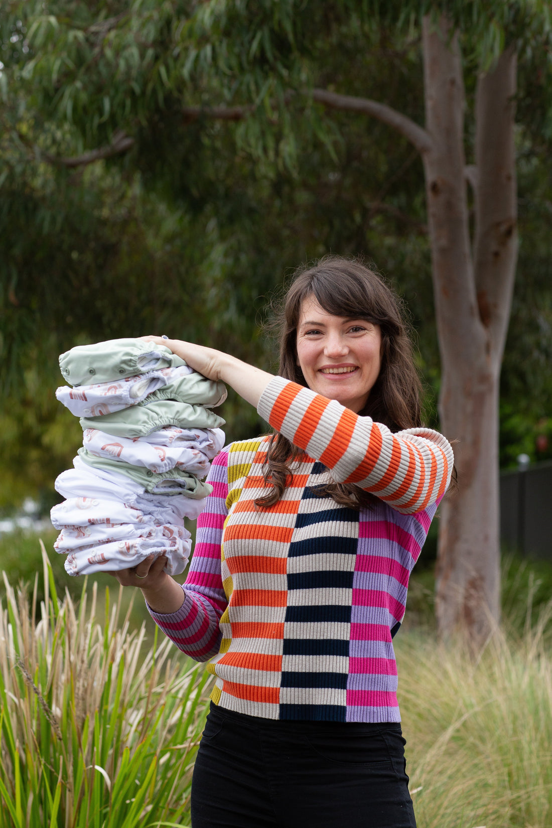 Woman holding a pile of cloth nappies in green and rainbow colours. Tree and grasslands are in the background.